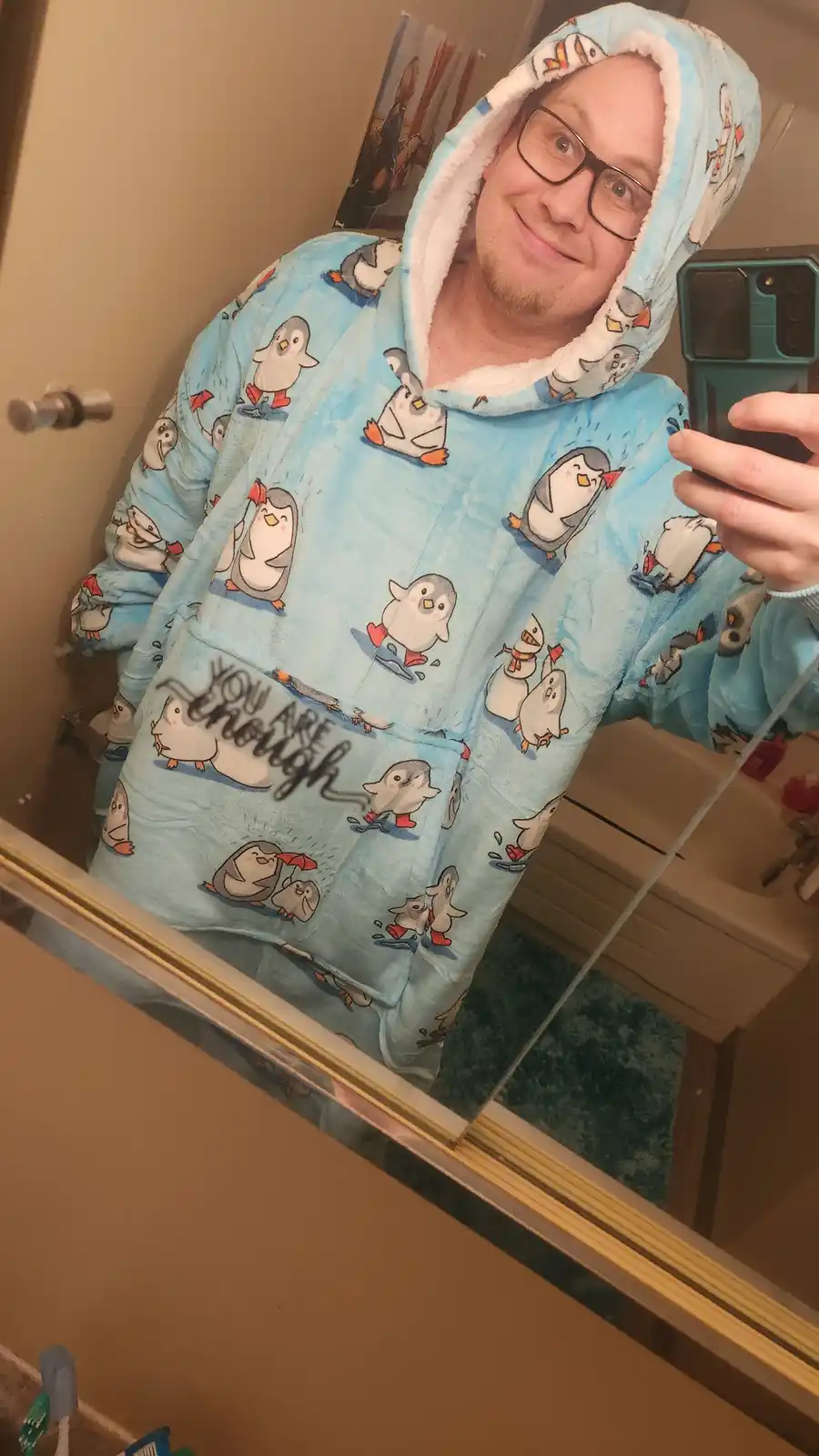 A person wearing a light blue, fluffy hooded robe covered in cartoon penguins takes a mirror selfie in a bathroom. The…