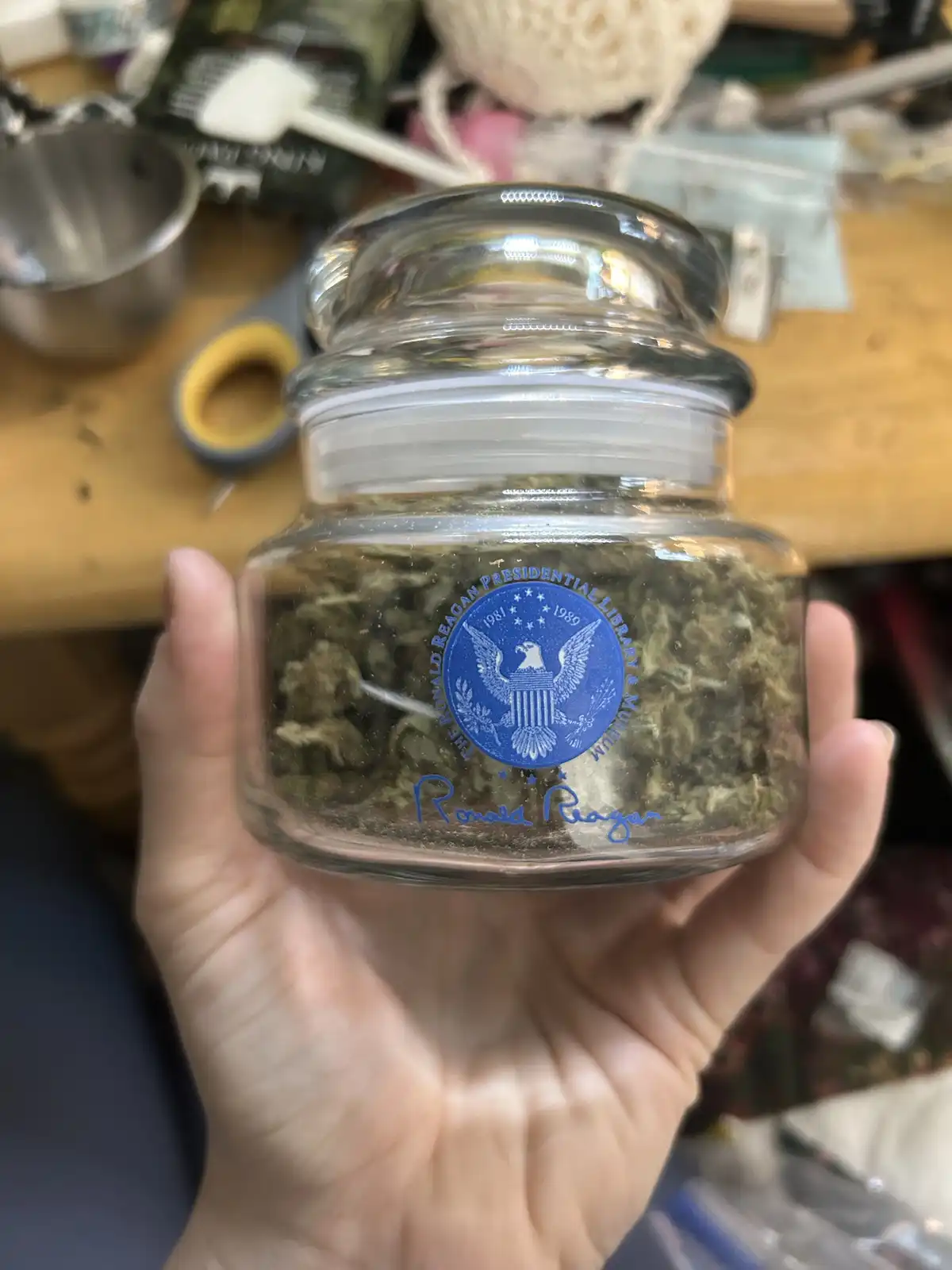 A hand holds a clear glass jar filled with green cannabis buds, which has a blue seal of the Ronald Reagan Presidential…