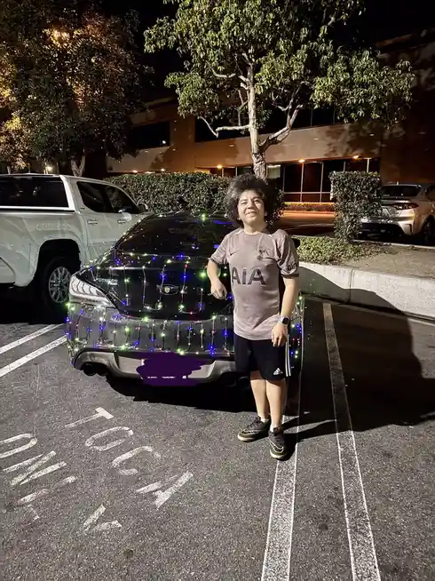 A person with curly hair stands proudly next to a dark-colored Genesis car parked at night in a lot, with colorful string…