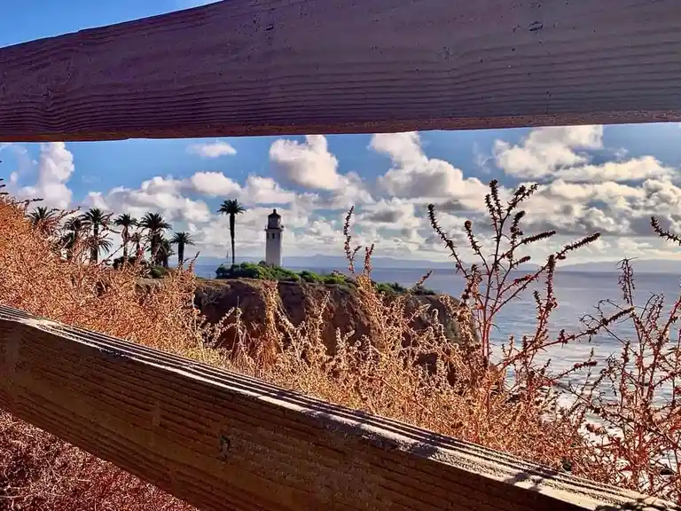 A scenic photo of a lighthouse on a cliff overlooking the ocean, framed by wooden fence rails in the foreground and dry…
