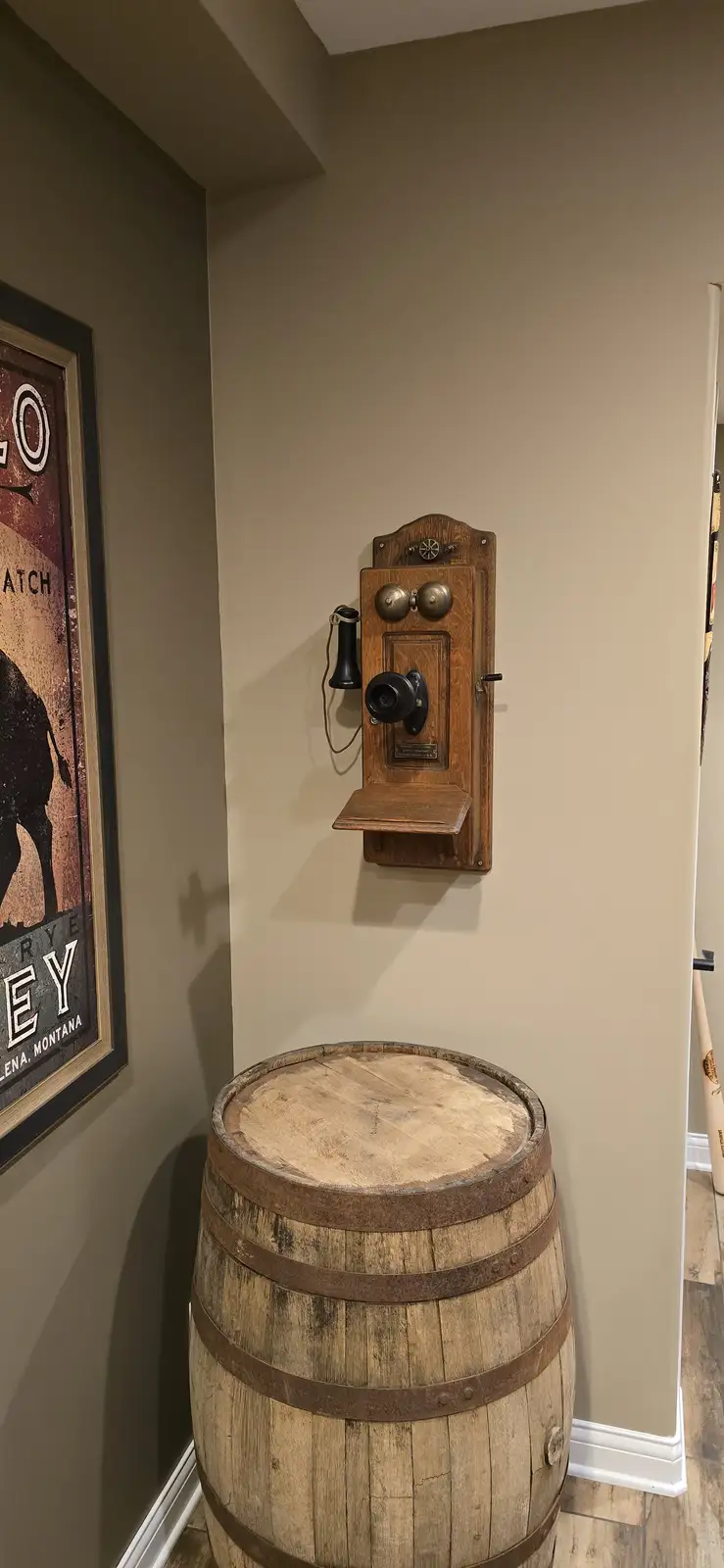 An antique wooden wall-mounted telephone with a black handset and dial is mounted above a large wooden barrel with metal…
