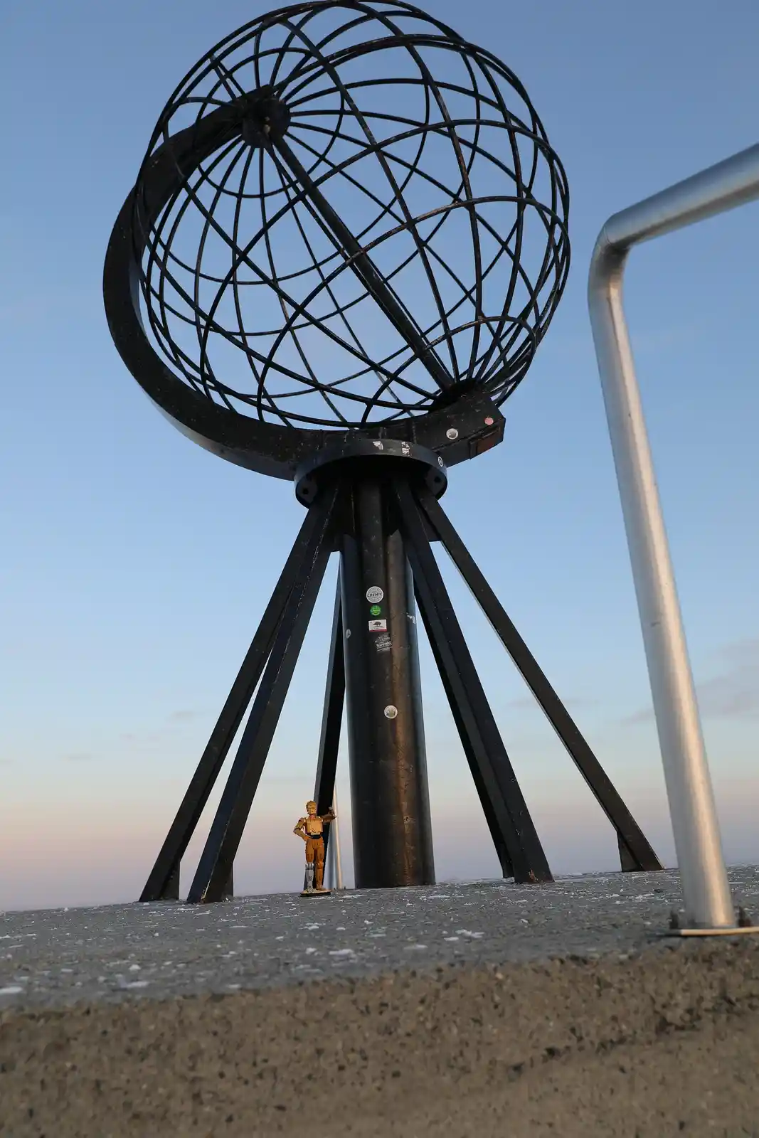 A small golden C-3PO action figure stands on a concrete surface next to a large black metal globe sculpture with a tripod…