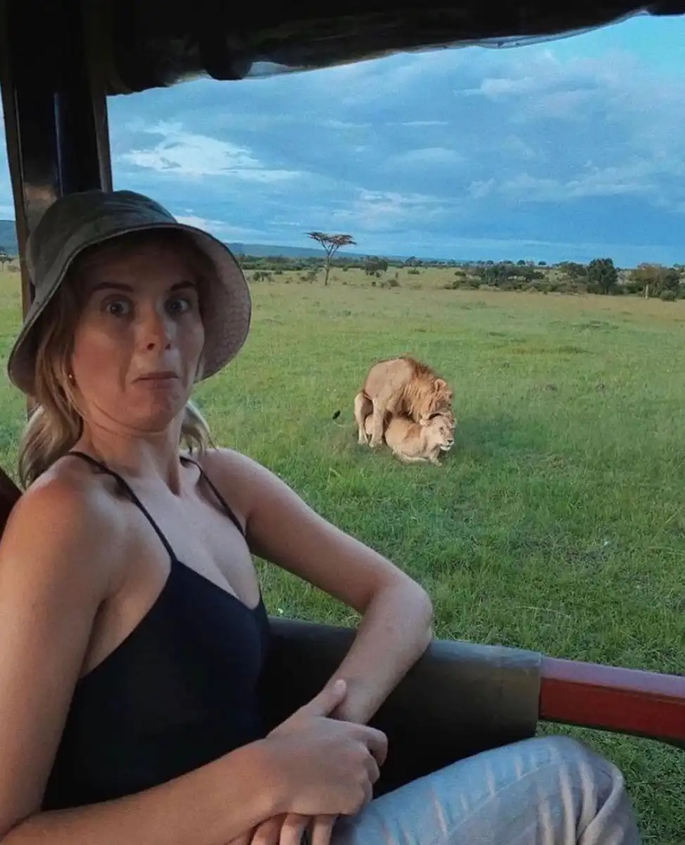 A woman wearing a bucket hat and black top sits in a safari vehicle, looking shocked at the camera. In the background, two…