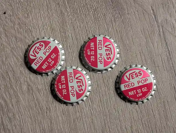 Four vintage Vess Red Pop bottle caps with red and white branding lie on a light wood-grain surface. The caps show 'VESS RED…