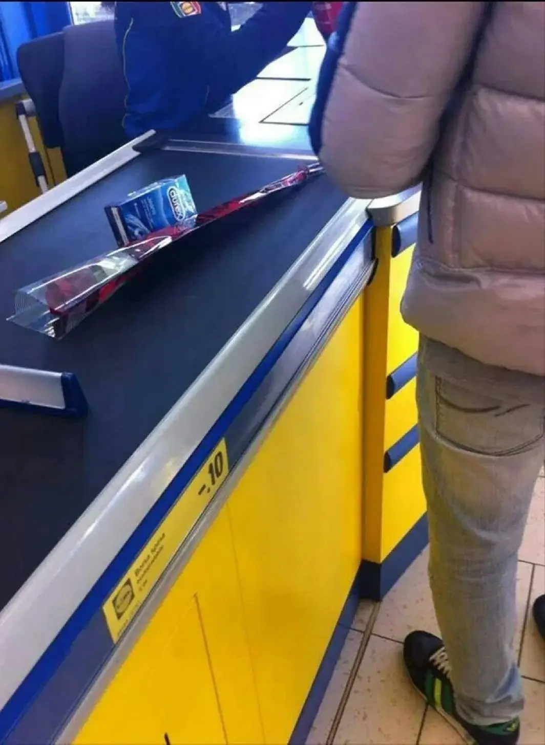 A person in a puffy jacket stands at a yellow and blue checkout counter in a store, with a packaged ski and a box of Oral-B…