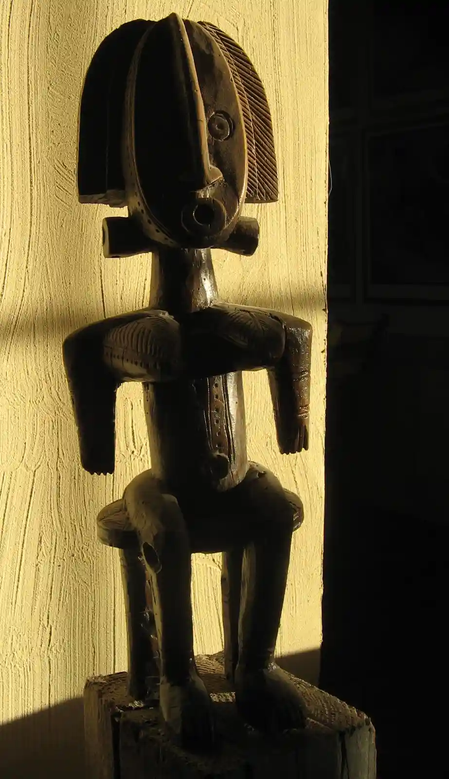 A dark wooden tribal statue with a stylized face, seated on a wooden base, is illuminated by a single spotlight against a…