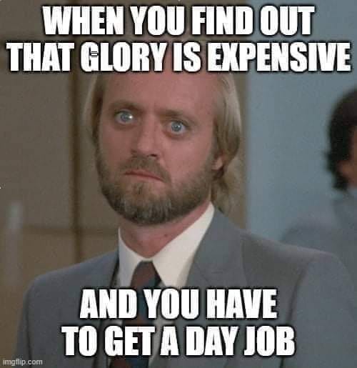 A man with blonde hair, a beard, and wide blue eyes stares with a shocked and dismayed expression while wearing a grey suit…