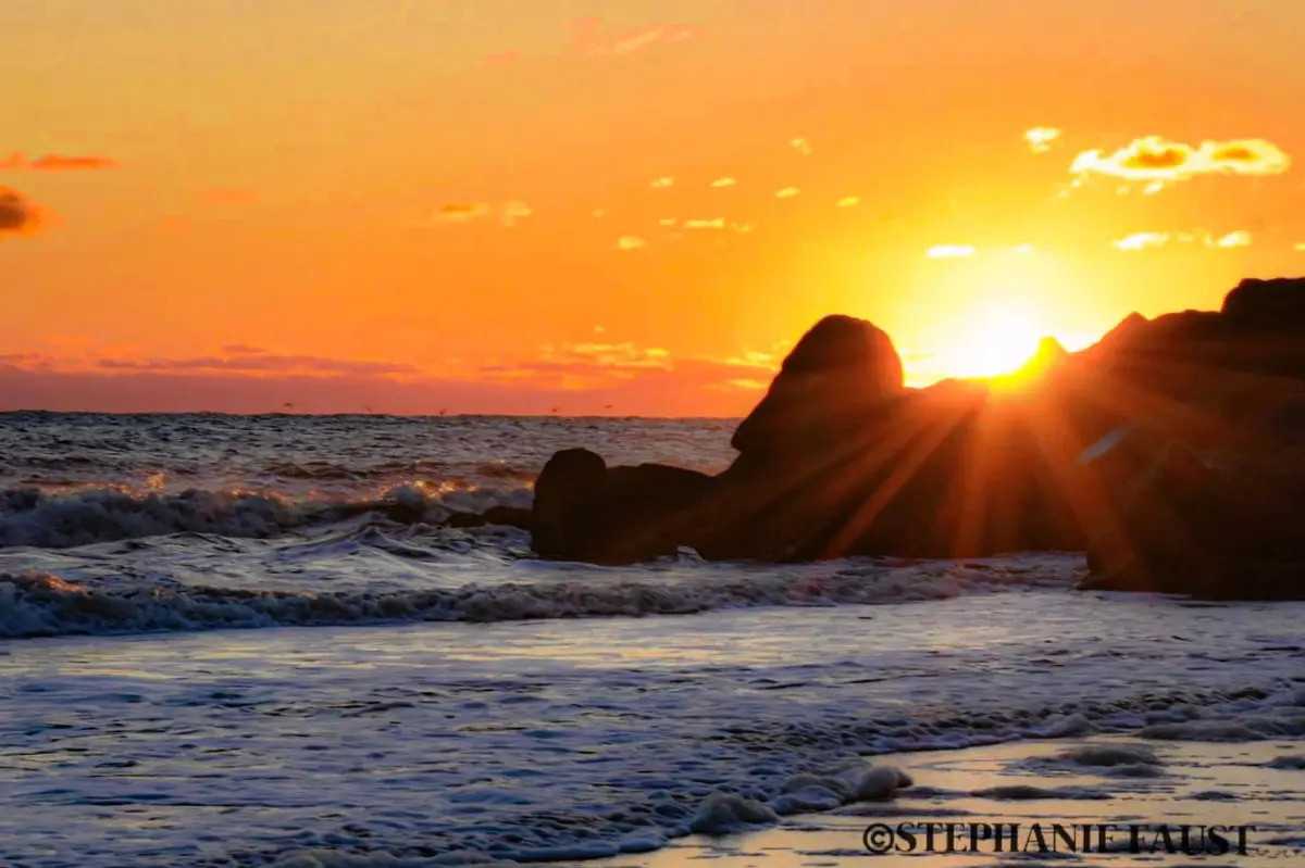A serene sunset over a rocky beach with waves crashing, the sun setting behind large rocks casting rays of light. The…