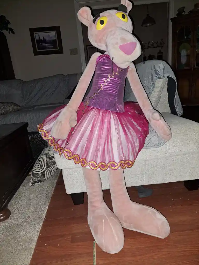 A large plush pink panther toy is dressed in a shiny pink and magenta ballet outfit with gold trim, sitting on a…