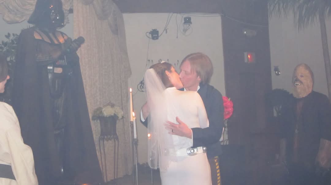 A bride and groom kiss during a wedding ceremony, with Darth Vader standing solemnly to the left and Chewbacca smiling to…