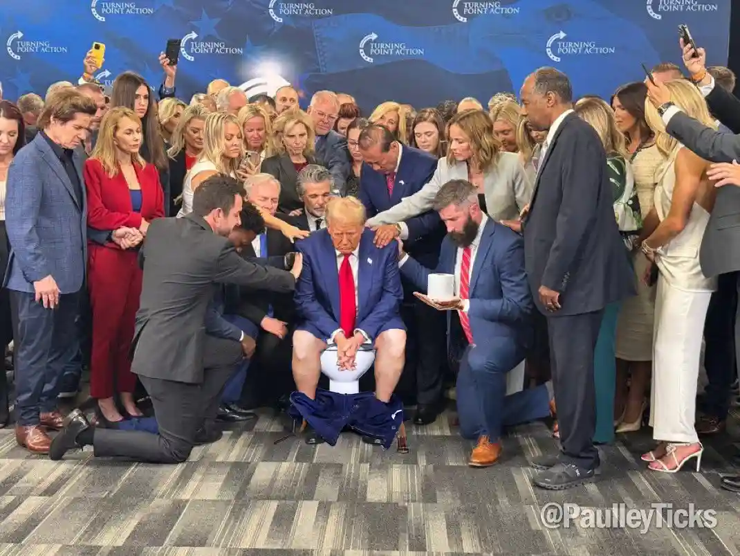 A man in a blue suit and red tie is seated on a white toilet in the middle of a crowded room, surrounded by people in…