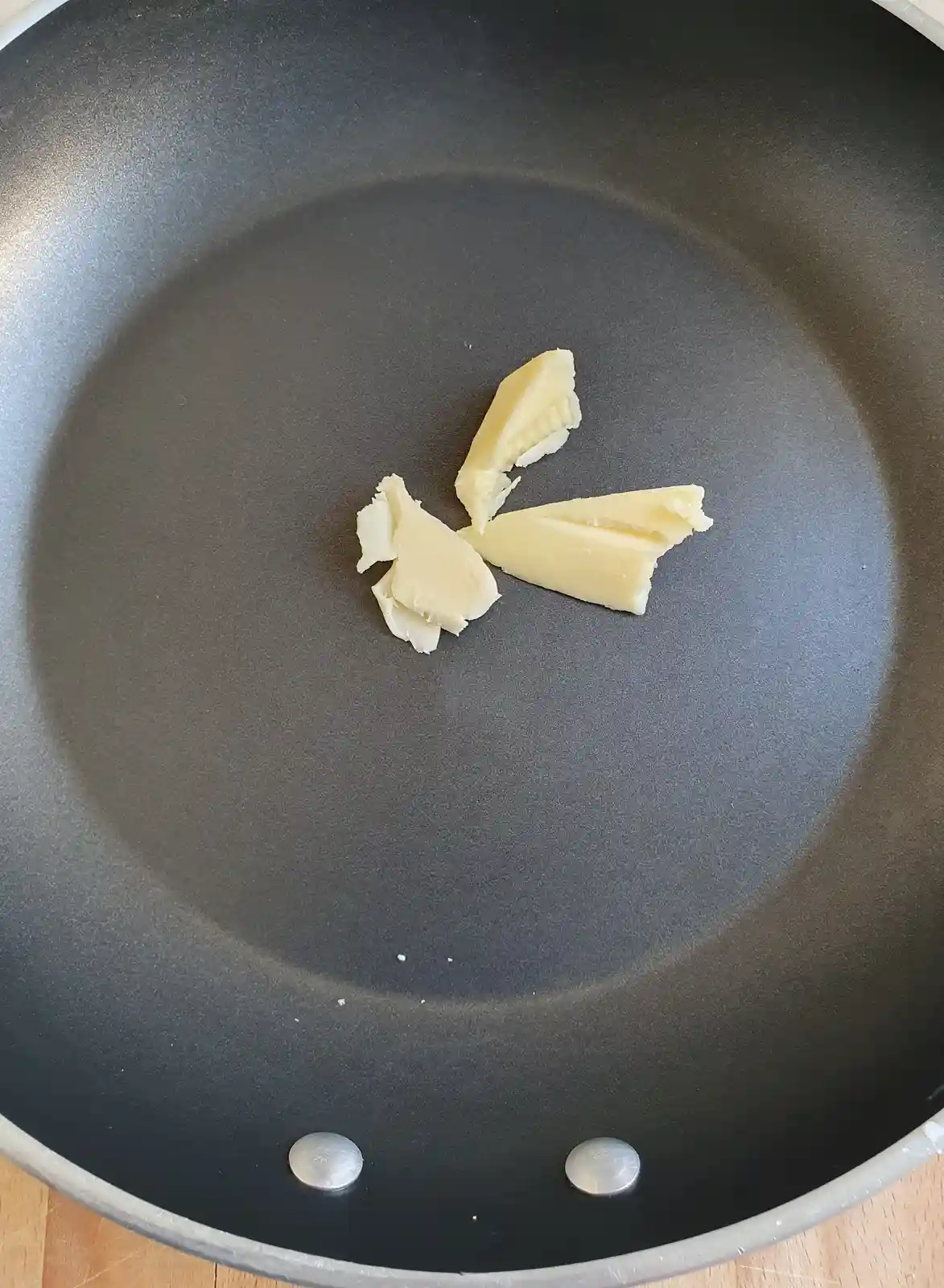 A black non-stick frying pan contains several irregularly shaped chunks of pale yellow butter arranged in a loose cluster…