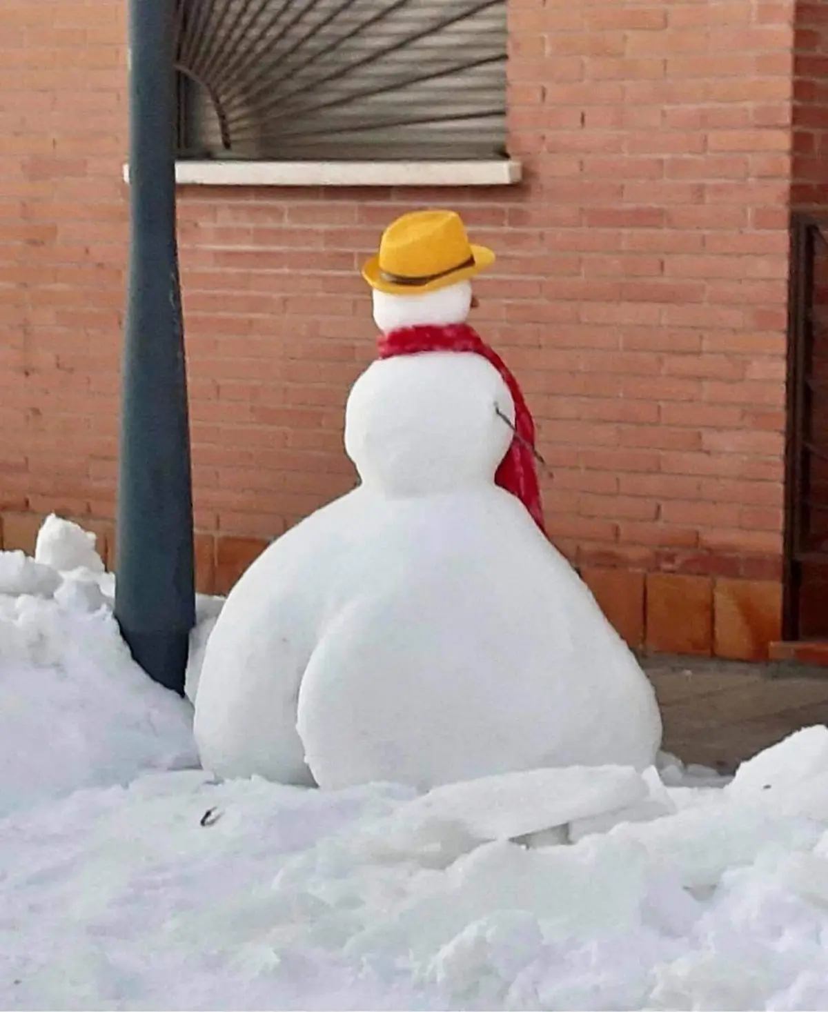 A snowman viewed from behind, wearing a yellow hat and red scarf, sits in a pile of snow in front of a brick building. The…