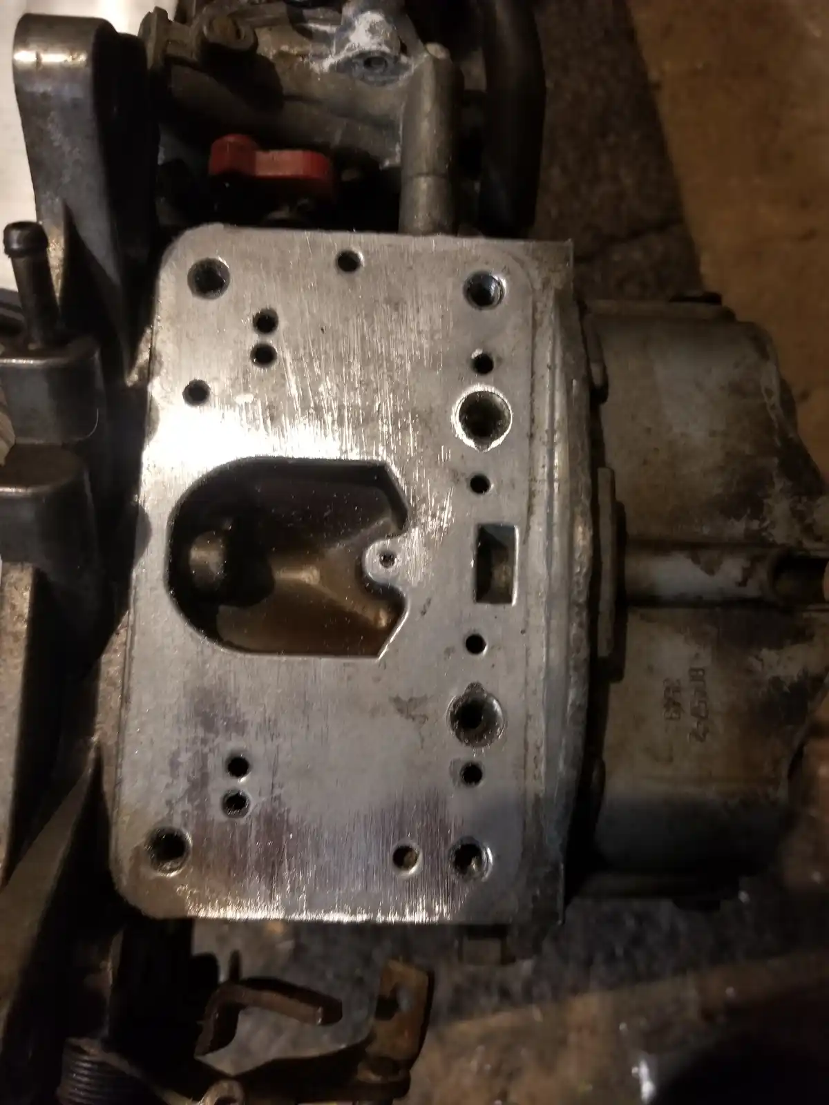 A close-up photo shows a dirty, metallic carburetor with visible bolts, ports, and a red lever, resting on a concrete floor…