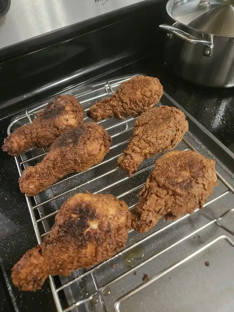 Six pieces of fried chicken, some with burnt spots, rest on a metal cooling rack atop a black stovetop. A stainless steel…