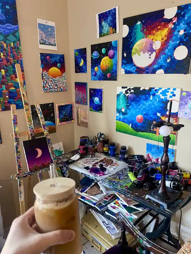 A hand holds an iced coffee in the foreground, with a cluttered artist's studio behind it. The walls are covered with…