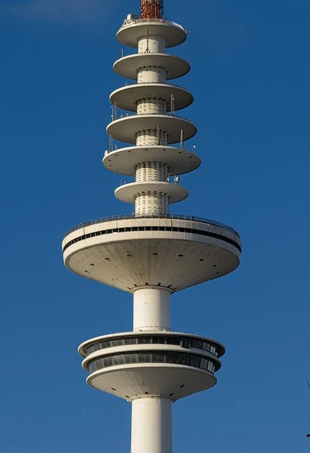 A tall, white, tiered observation tower with circular platforms against a clear blue sky. The image has overlaid text…