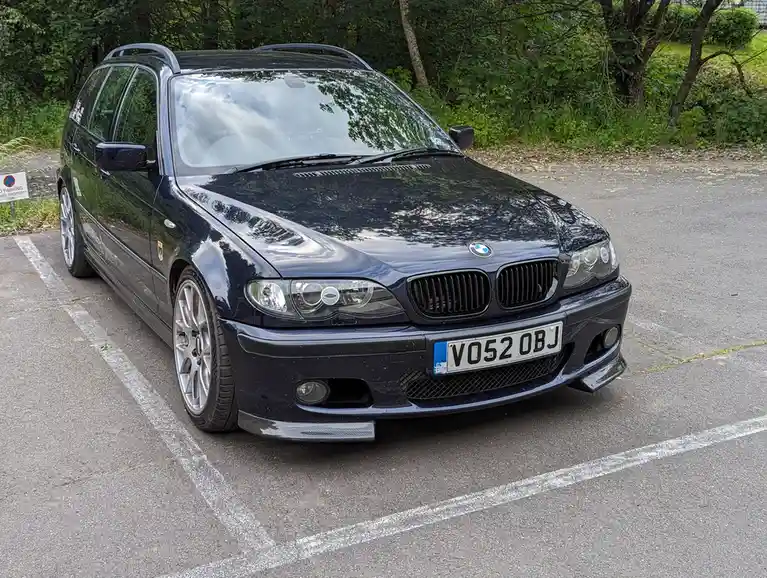 A dark blue BMW 3-Series Touring station wagon is parked in an asphalt lot with white parking lines, surrounded by green…