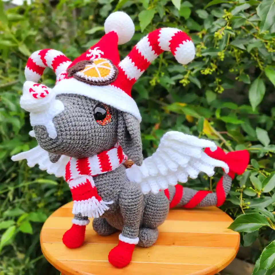 A gray crocheted amigurumi dog is dressed in festive holiday attire, including a red and white jester hat with candy cane…