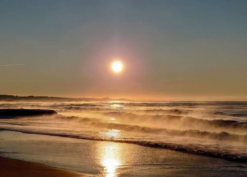 A serene beach scene at sunrise or sunset with the sun low in the sky, casting golden light across misty ocean waves rolling…