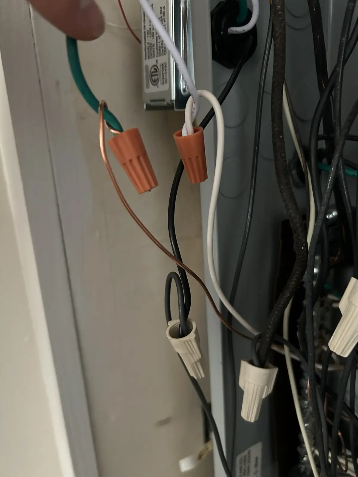 A close-up photo shows a person's finger holding a green wire connected to an orange wire nut, with other wires and…