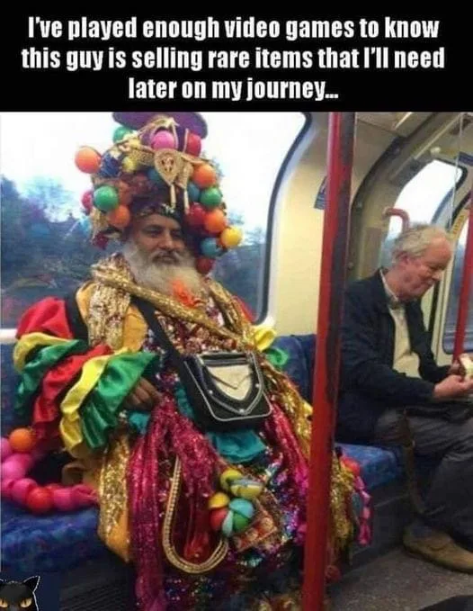 A man sits on a public transit train wearing an elaborate, colorful costume with a large, multi-colored hat, gold chains…