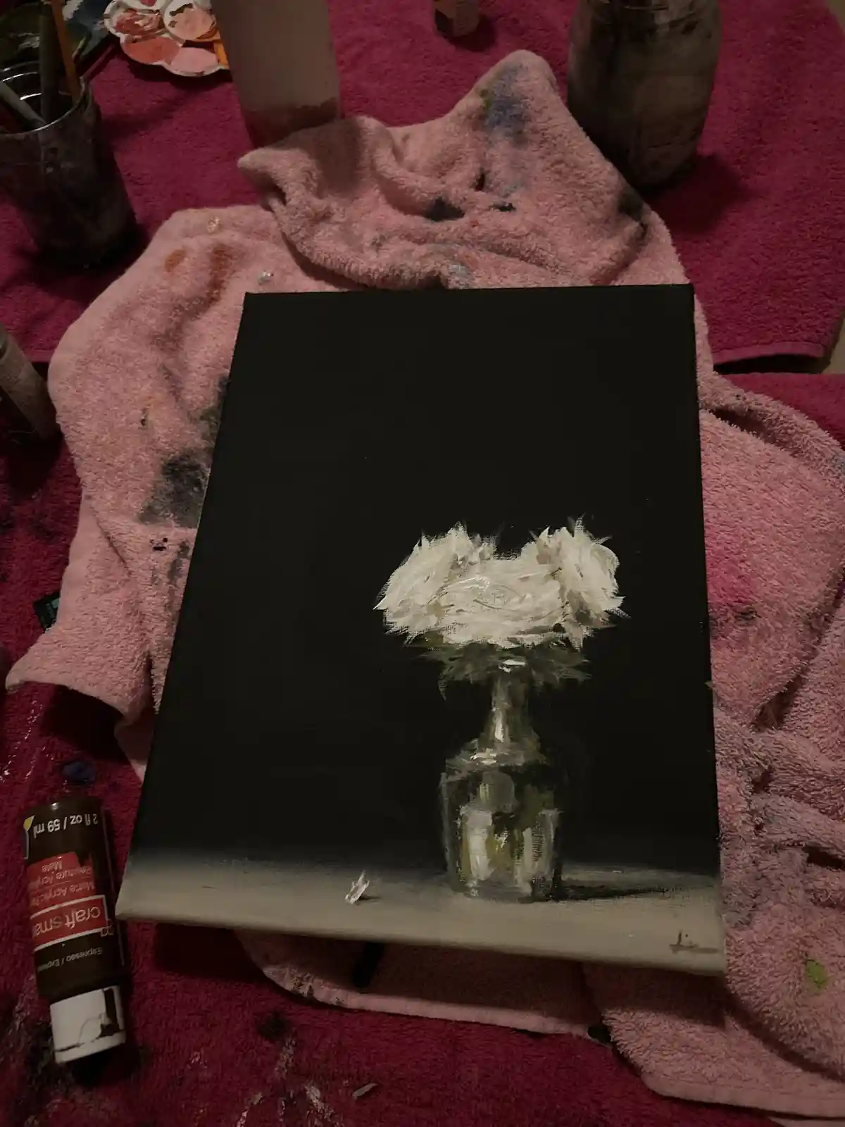 A painting of white roses in a glass vase on a black background sits on a pink towel amidst paint supplies. The overlaid…