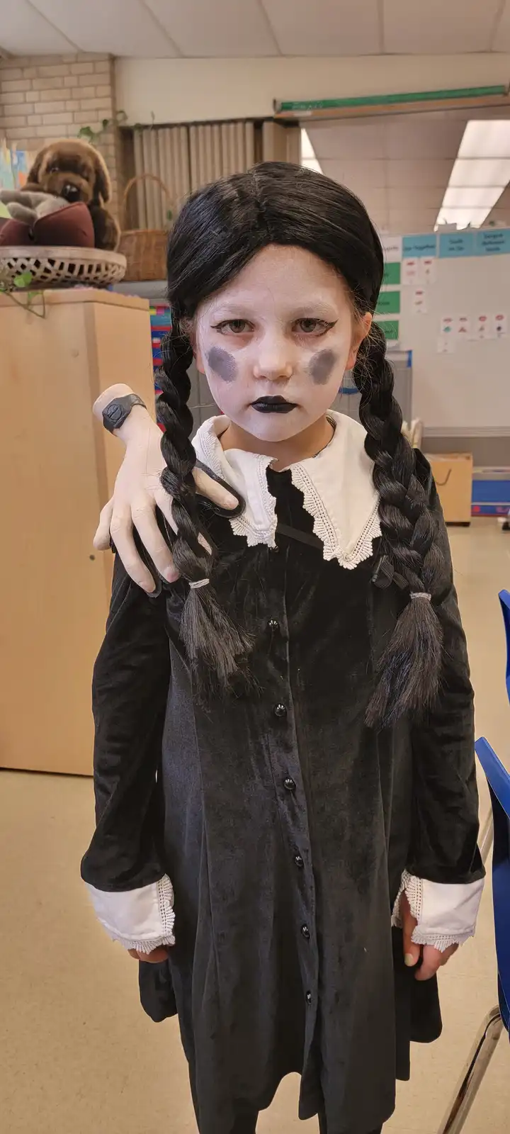 A young child dressed as Wednesday Addams, complete with black dress, white lace collar, black braids, pale face paint, dark…