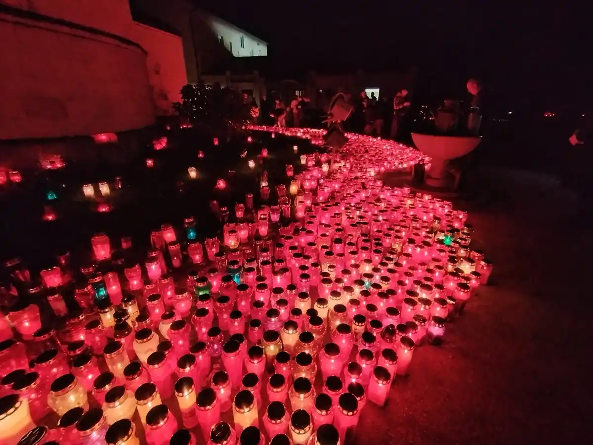 A nighttime photo shows hundreds of lit candles in jars arranged in a large, glowing pattern on the ground, mostly pink and…