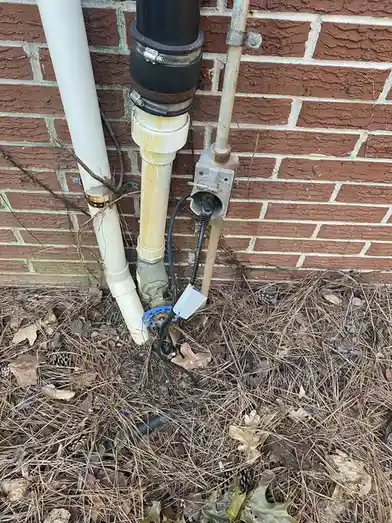A close-up photo shows outdoor plumbing pipes against a brick wall, with a black power cord plugged into a metal junction…