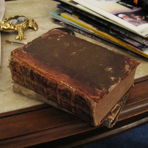 A heavily worn, brown leather-bound book sits on a wooden table, its cover and spine visibly aged and damaged. Behind it, a…