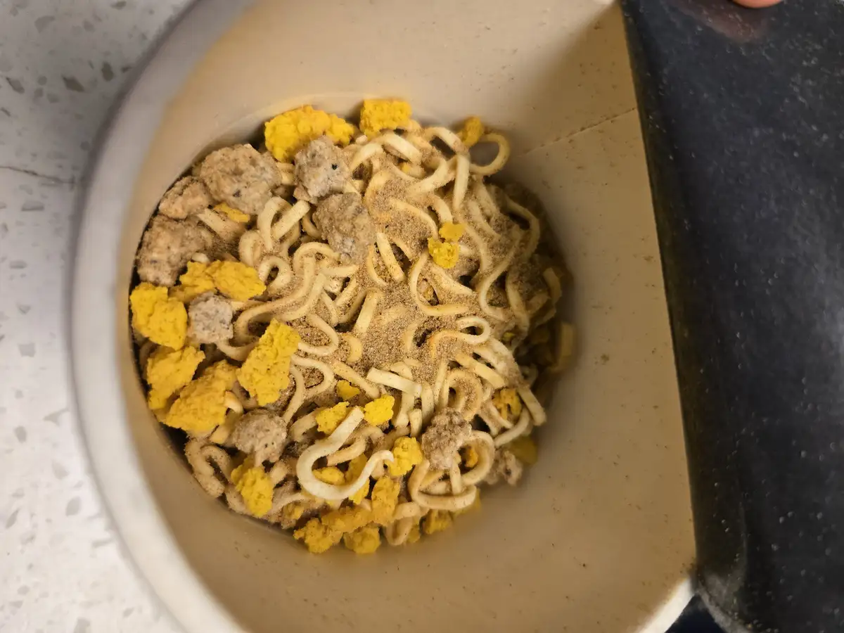 A top-down view of a beige ramen cup filled with uncooked noodles, yellow egg chunks, and brown meatballs, all heavily…