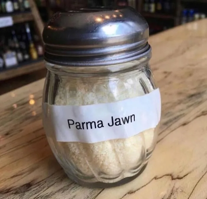 A clear glass shaker filled with grated cheese sits on a wooden table, labeled with a white paper sticker that reads 'Parma…