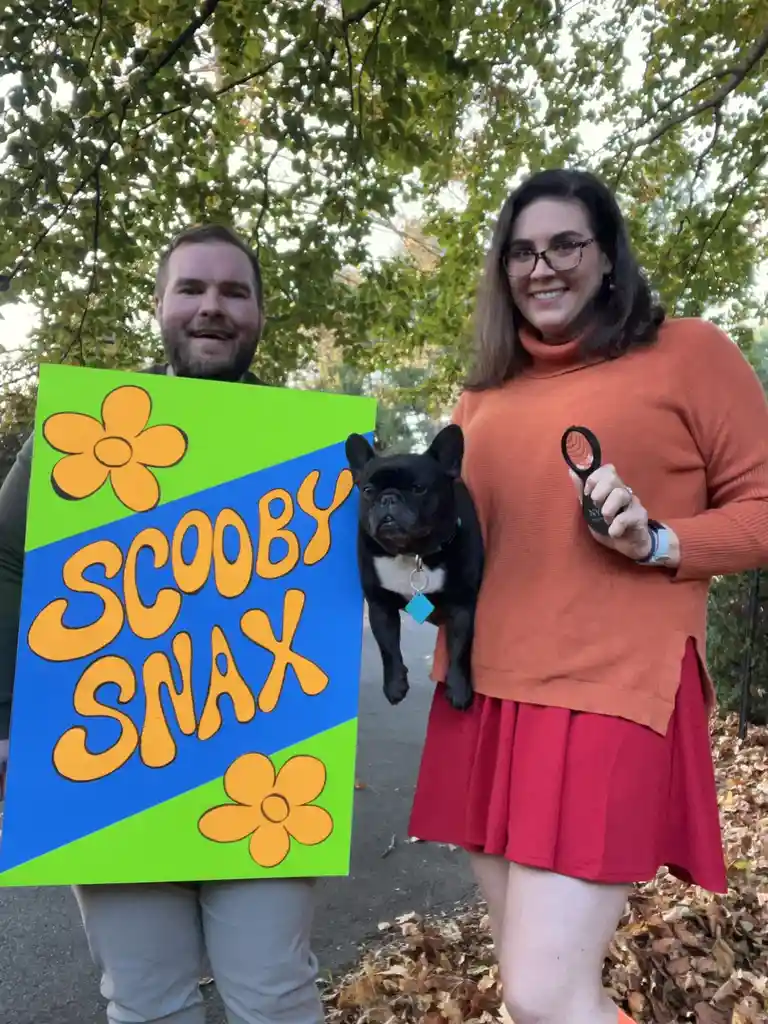 Two people dressed as Scooby-Doo and Shaggy stand outdoors holding a large sign that reads 'SCOOBY SNAX' with orange flowers…
