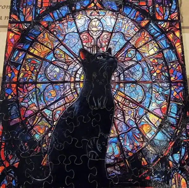 A completed jigsaw puzzle depicts a black cat sitting upright against a colorful stained-glass window background, with the…