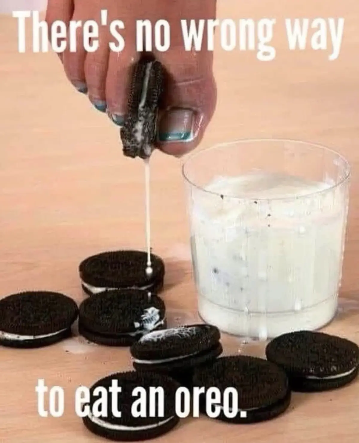 A hand with blue-tipped fingernails holds an Oreo cookie, squeezing the cream filling directly into a glass of milk. Several…