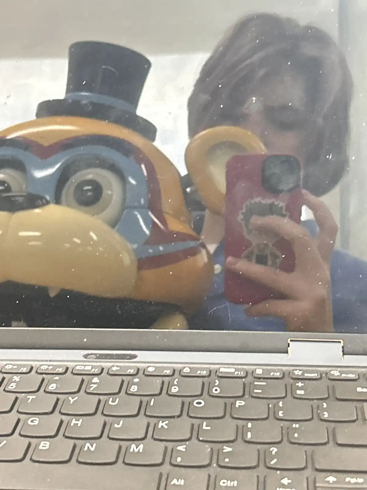 A mirror selfie shows a person holding a red phone, standing beside a large, yellow animatronic figure wearing a top hat…