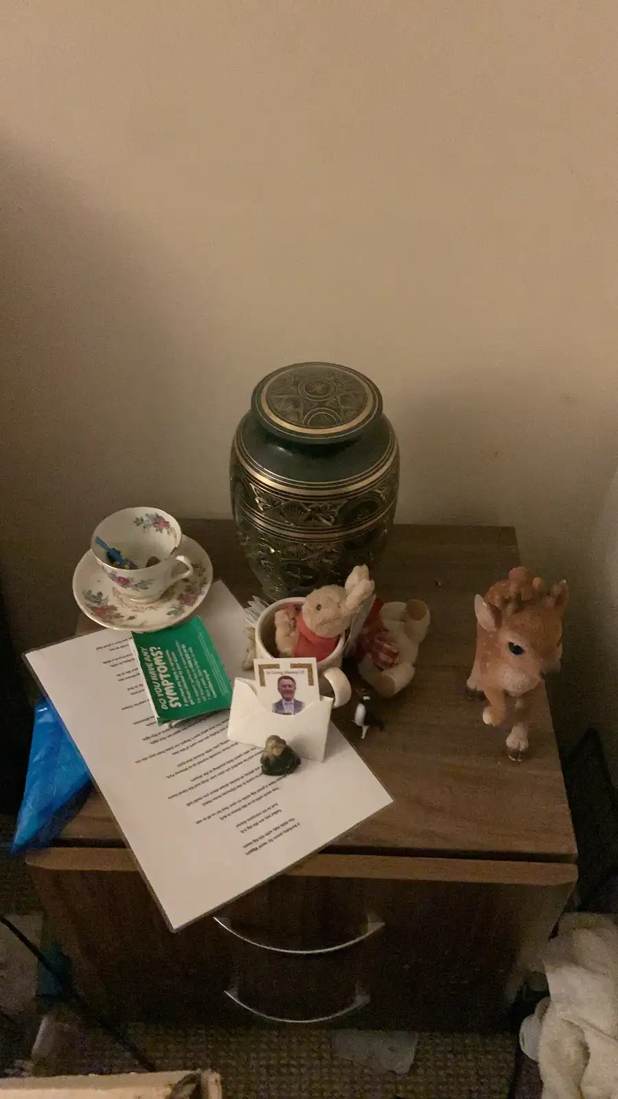 A cluttered bedside table holds a green urn, a floral teacup, stuffed animals, a plastic deer figurine, a document titled…