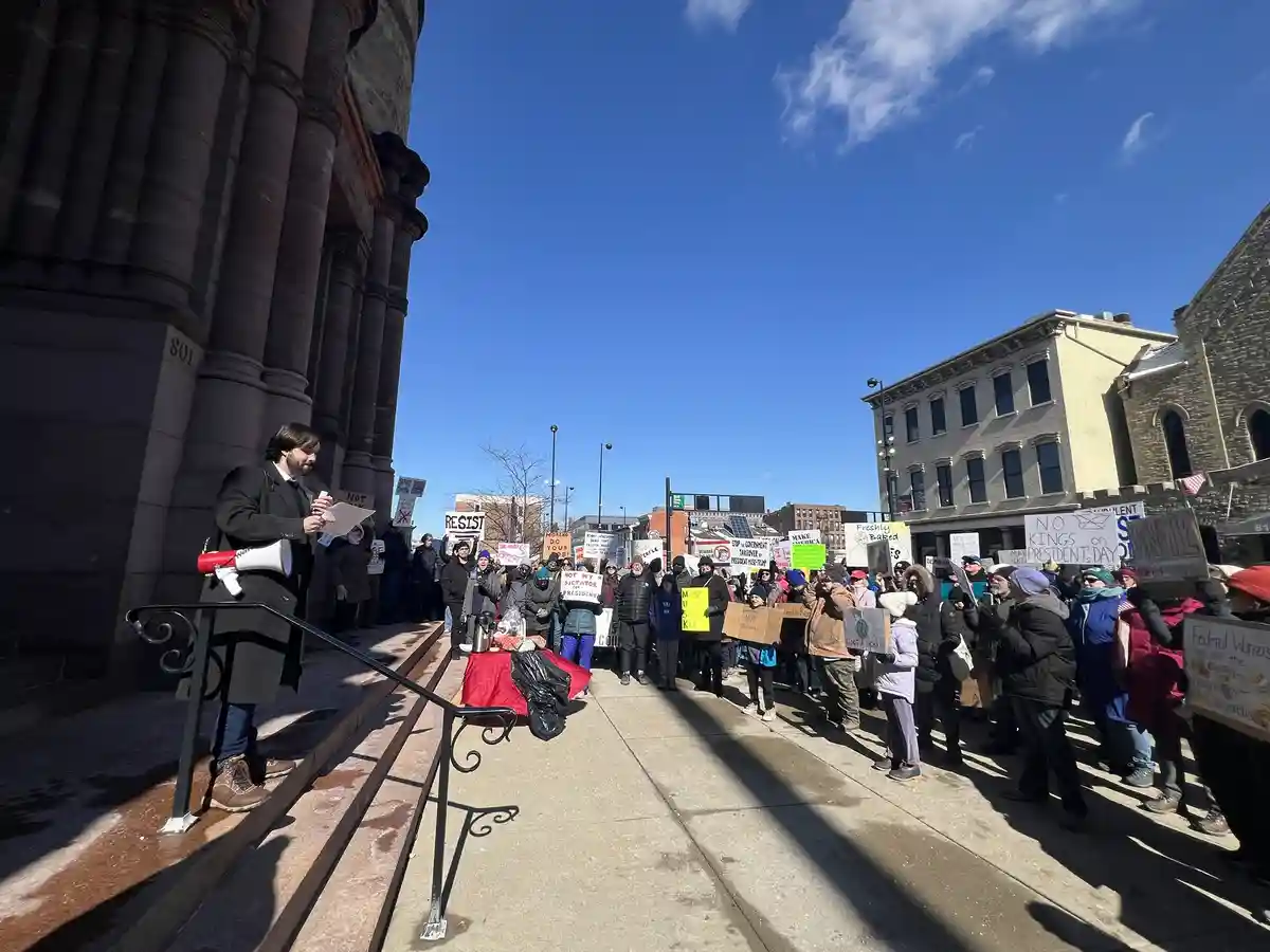 A man in a black coat stands on steps holding a megaphone and papers, addressing a crowd of protesters holding signs like…