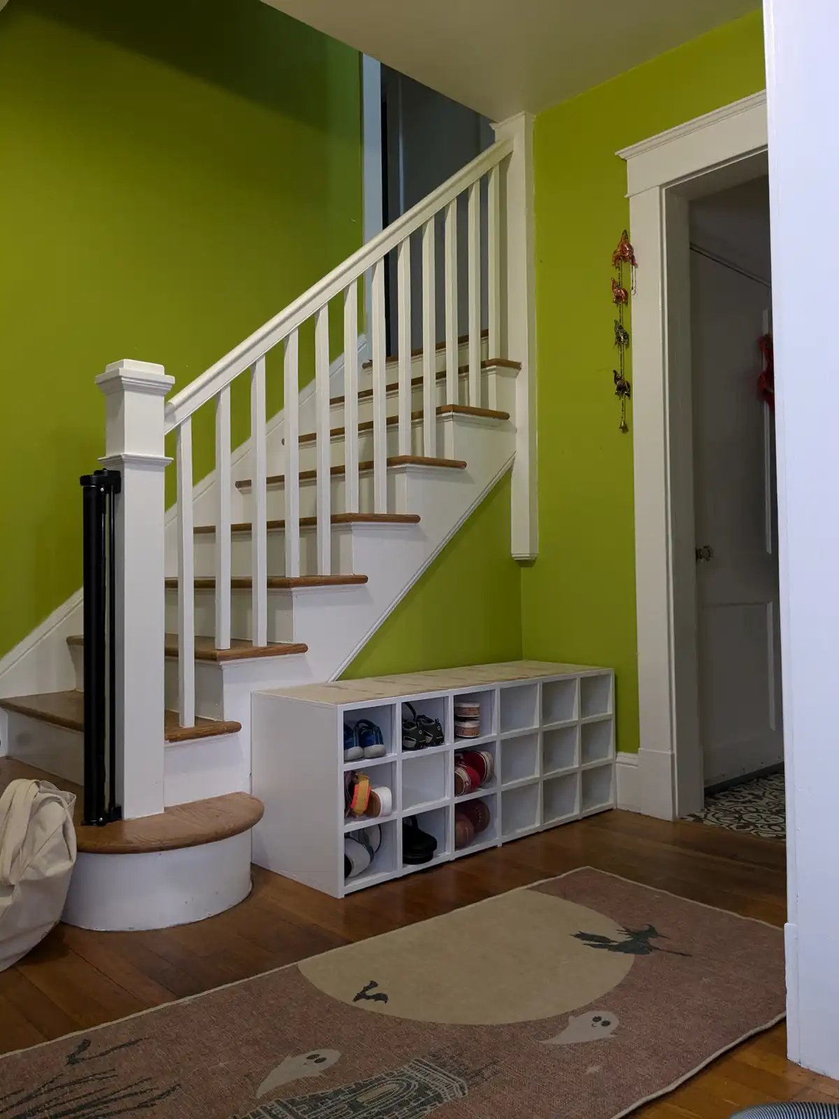 A white, cube-style shoe rack sits at the base of a staircase in a home with lime green walls and hardwood floors. The rack…