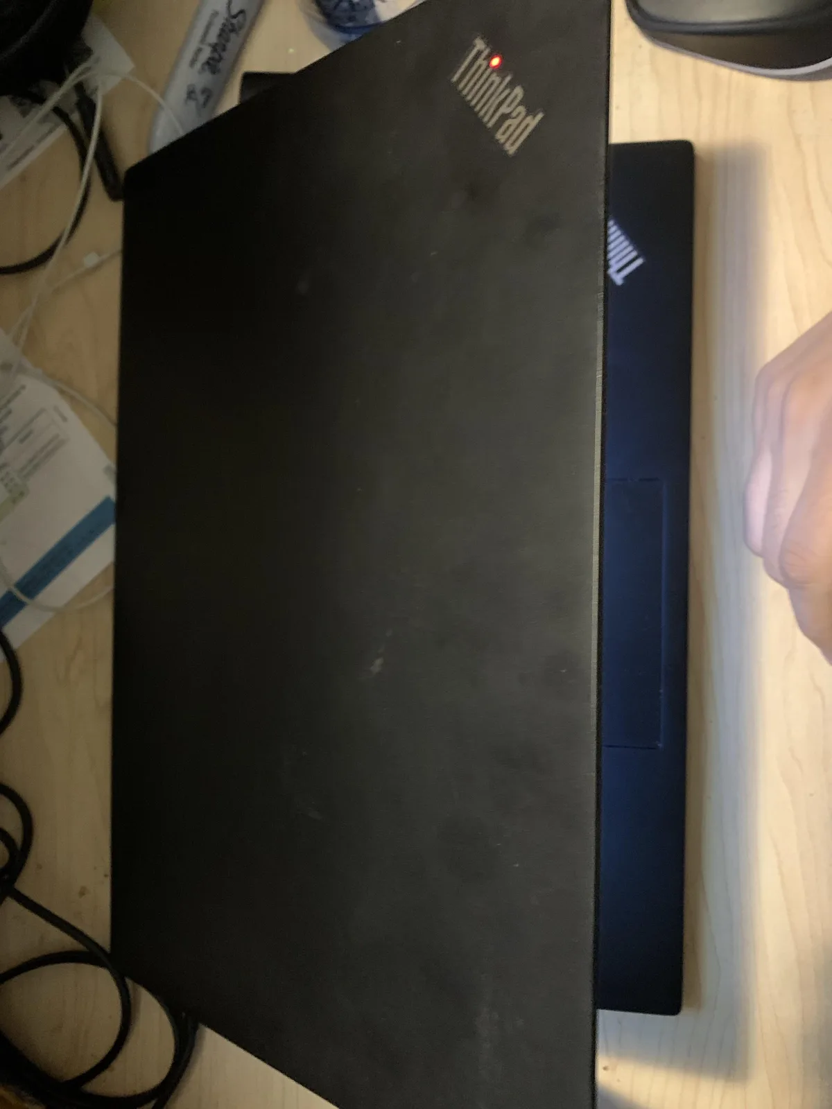 A black Lenovo ThinkPad laptop is shown closed on a wooden desk, with its logo and a red power indicator light visible on…