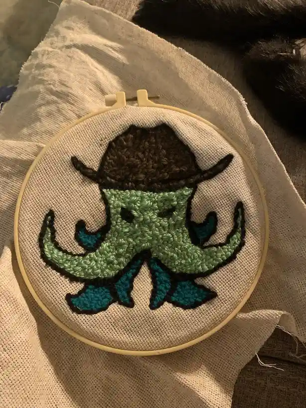 A close-up photo shows a hand-embroidered design of a green octopus wearing a brown hat and blue boots, stitched onto…