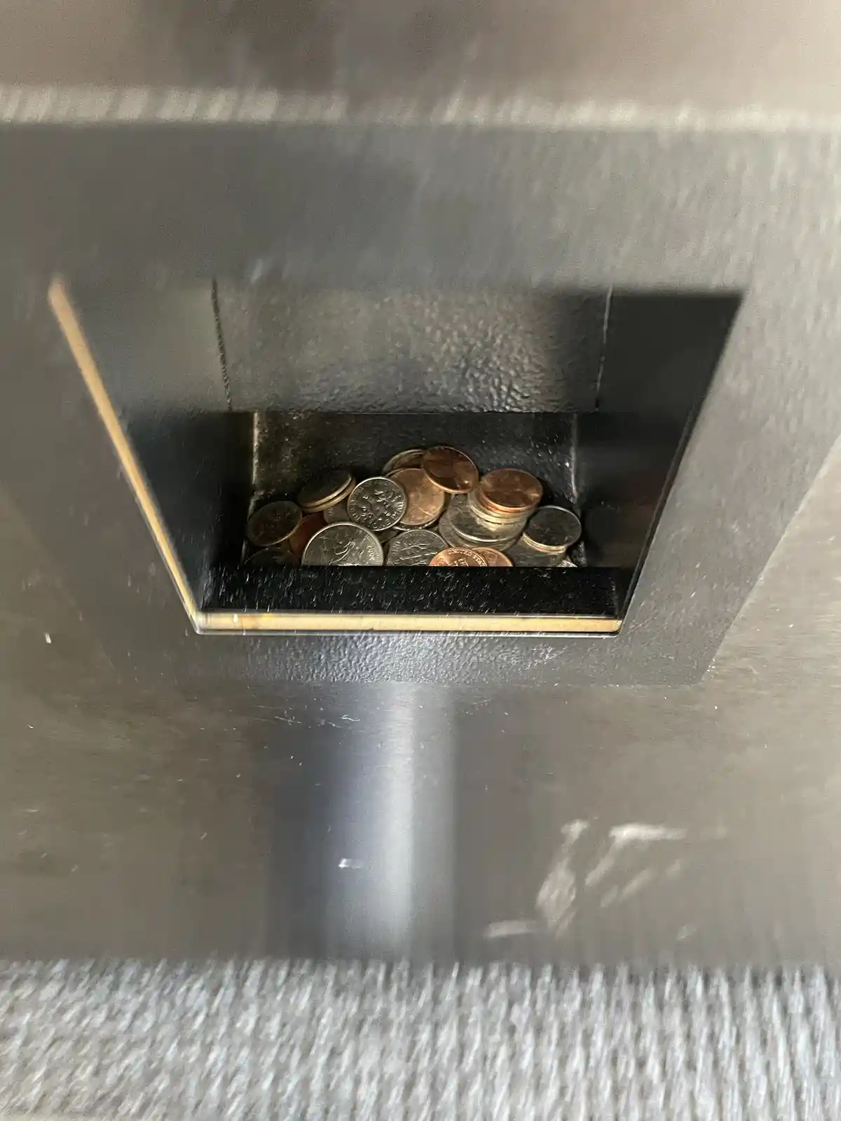 A close-up shot shows a dark, textured coin slot of a vending machine filled with a mix of copper and silver coins. The slot…