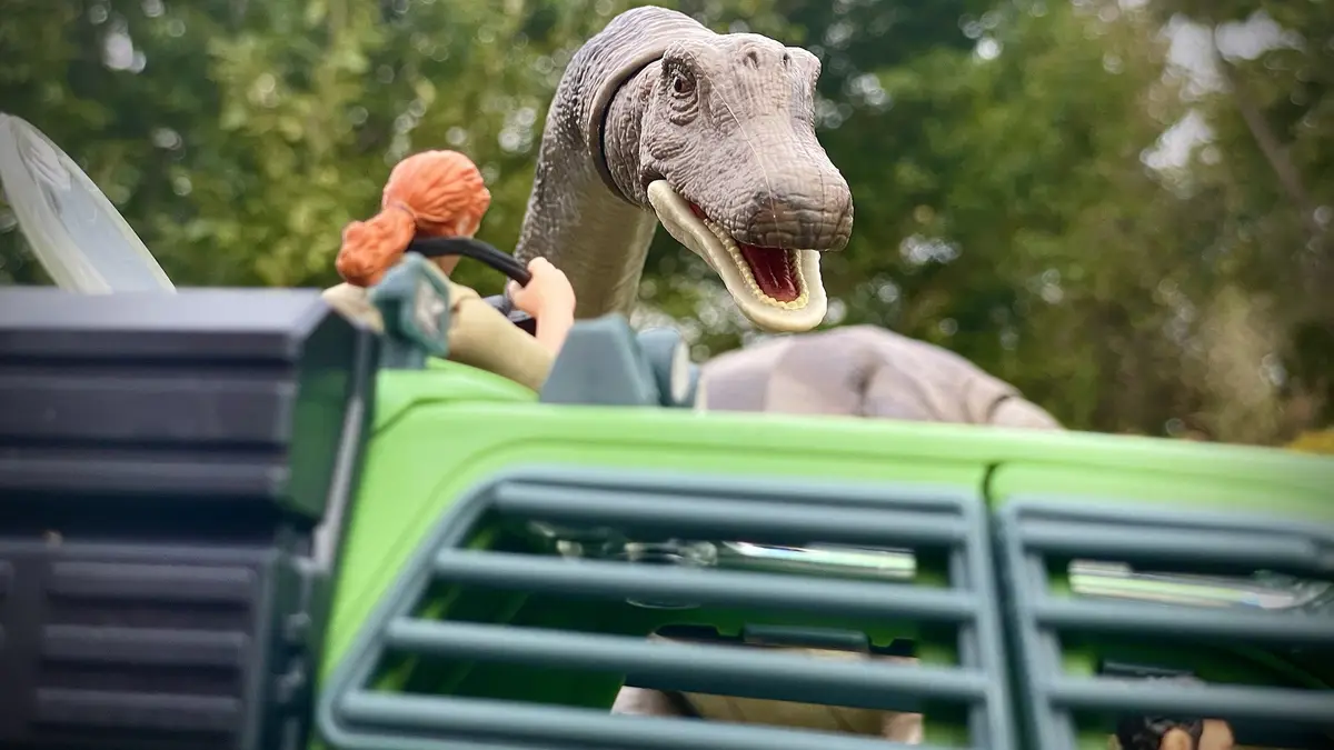 A close-up shot of a toy dinosaur with its mouth open, positioned behind a green toy vehicle. A small toy figure with orange…