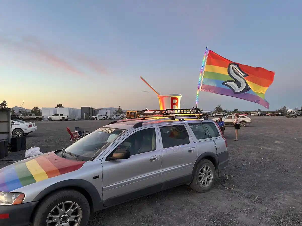 A silver station wagon is parked in a gravel lot at dusk, decorated with a rainbow flag on its roof rack and a rainbow…