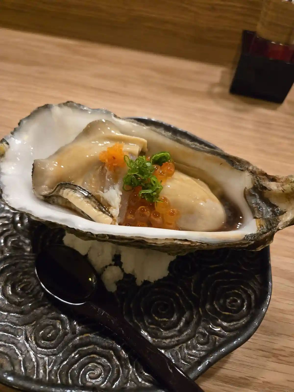 A raw oyster served on the half shell, garnished with orange tobiko (fish roe) and green chopped herbs, sits atop a bed of…