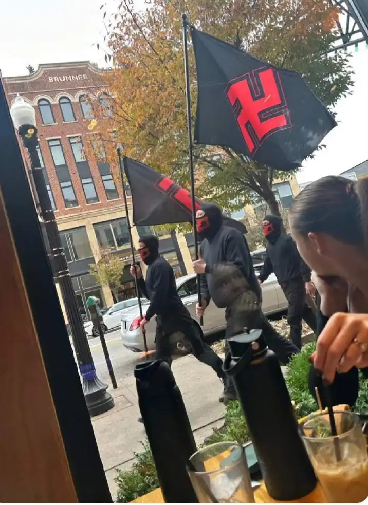 A photo taken from inside a cafe shows people outside holding black flags with red swastikas, while a person inside looks…