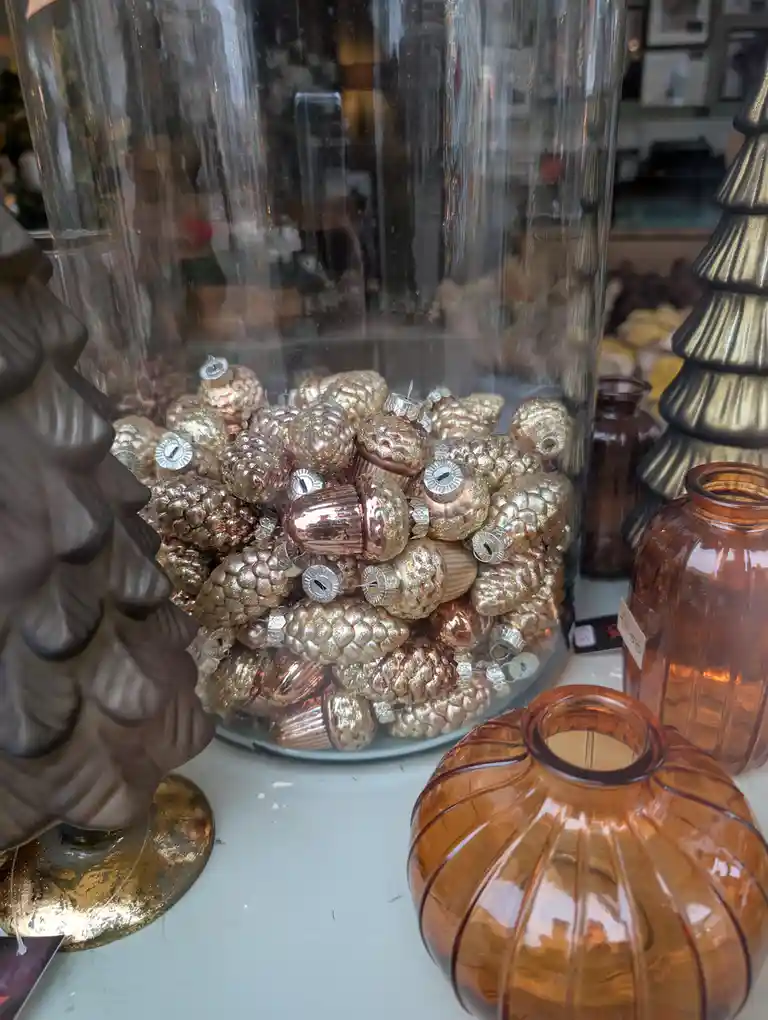 A large glass jar filled with shiny, metallic gold pinecone-shaped Christmas ornaments sits on a display shelf, surrounded…
