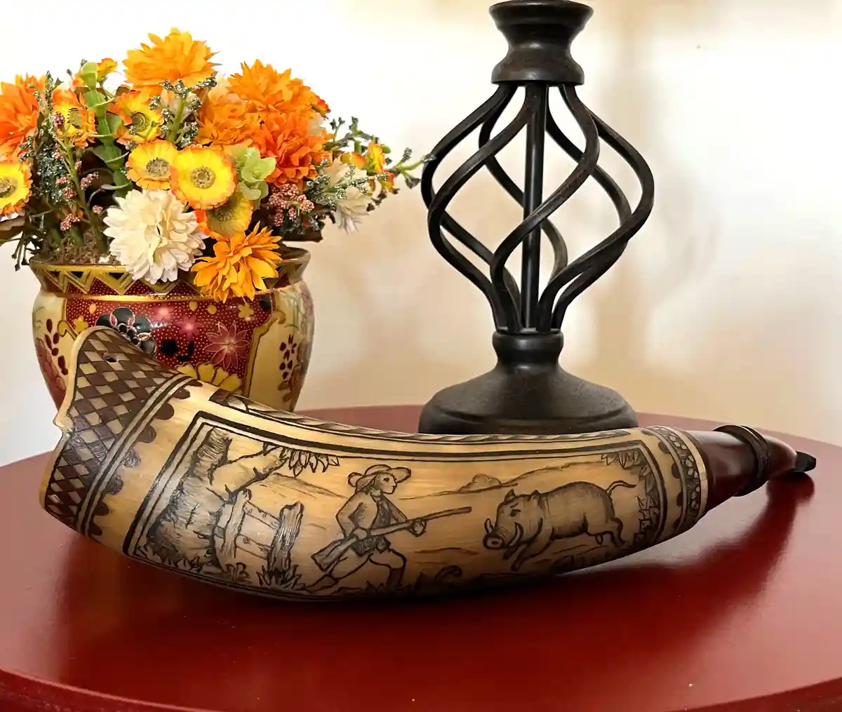 A carved wooden hunting horn with a scene of a hunter aiming at a wild boar is displayed on a red table next to a vase of…
