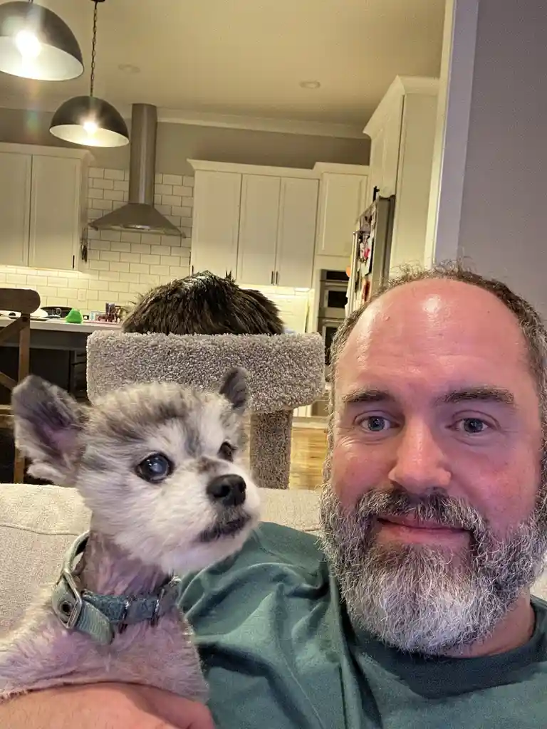 A man with a gray beard and blue eyes smiles at the camera while holding a small, shaved husky puppy with blue eyes and a…